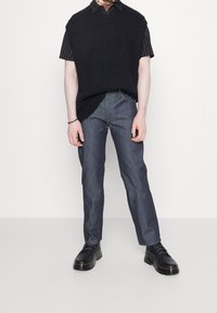 Svart stickad ärmlös tröja över en randig kortärmad skjorta, parat med mörk denimjeans och svarta snörstövlar. Minimalistisk design.