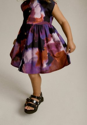 Une robe multicolore présentant des motifs aquarelle en violet, rouge et blanc, avec un design à boutons à l'avant, associée à des sandales noires.