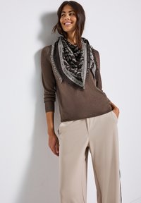 Brun långärmad tröja i kombination med ljusbeige byxor. En svart och gråmönstrad scarf med fransar är draperad runt halsen.