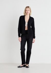 Cardigan negru cu textură în relief, cu deschidere în față, purtat peste un top alb, combinat cu blugi negri din denim cu accente cu cristale pe laterale și pantofi fără toc.