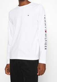 T-shirt bianca a maniche lunghe in cotone con colletto rotondo, piccolo logo sul petto e scritta "TOMMY HILFIGER" stampata verticalmente sulla manica.