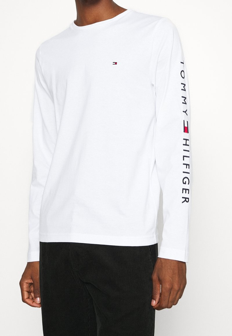 T-shirt bianca a maniche lunghe in cotone con colletto rotondo, piccolo logo sul petto e scritta "TOMMY HILFIGER" stampata verticalmente sulla manica.