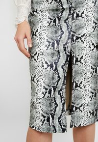 Kvinnas hand vilar på höften, klädd i en vit spetsärmad blus och en knälång svart och grå snakeskin-mönstrad kjol med en mittslits.