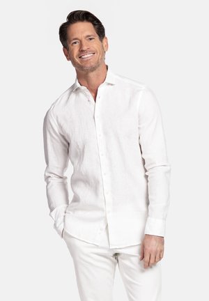 Glimlachende man met bruin haar en lichte stoppels, gekleed in een wit overhemd met lange mouwen en witte broek, voor een effen achtergrond.