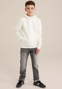 Sudadera de crema clara con un bolsillo frontal, combinada con jeans gris desgastados y zapatillas negras. Tela suave, corte relajado y capucha ajustable.