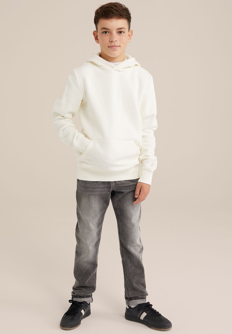Sudadera de crema clara con un bolsillo frontal, combinada con jeans gris desgastados y zapatillas negras. Tela suave, corte relajado y capucha ajustable.