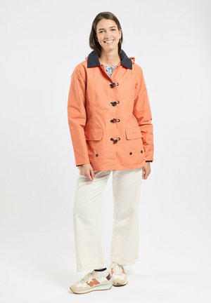 Femme souriante portant une veste à capuche orange avec des boutons à bascule, un pantalon blanc à jambes larges et des baskets beige avec des accents orange.