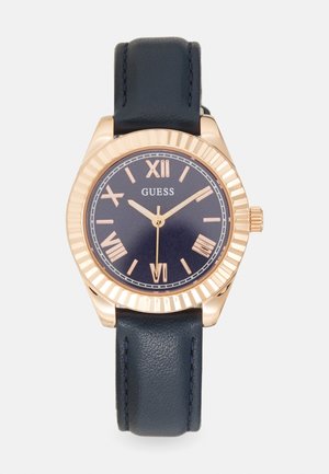 Rose guld ur med rem i marineblåt læder, rund urskive, romerske tal som markører og en tekstureret bezel. Brandnavnet "GUESS" vises.