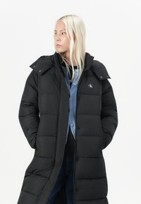 Svart pufferjakke med langt design, som har høy krage, trykknapper og logo. Laget av quiltet stoff, gir det et varmt utseende.