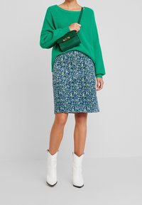 Jersey verde de punto, falda floral azul, botas de cowboy blancas. Pequeño bolso negro con detalle dorado, corte relajado, materiales texturizados.