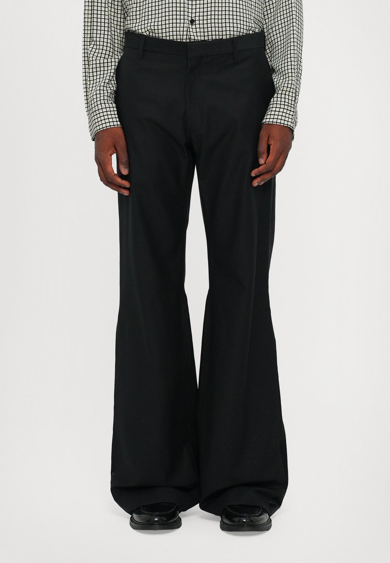 Pantalon noir à jambes larges avec une texture lisse, présentant une taille cintrée et une silhouette fluide, associé à des chaussures noires.