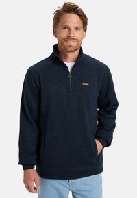 Navy fleece pullover with a quarter-zip, textured fabric, ribbed cuffs, and a small logo patch on the left chest. Front pockets visible.