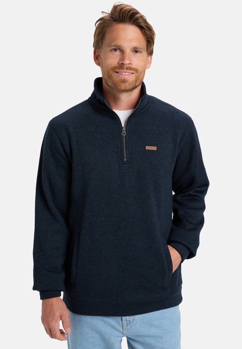 Navy fleece pullover with a quarter-zip, textured fabric, ribbed cuffs, and a small logo patch on the left chest. Front pockets visible.