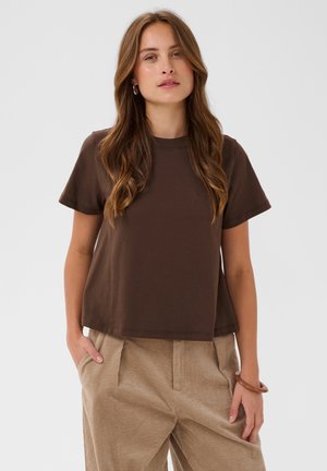 Femme aux longs cheveux bruns, portant un t-shirt marron foncé à manches courtes et un pantalon beige, debout les mains dans les poches devant un fond uni.