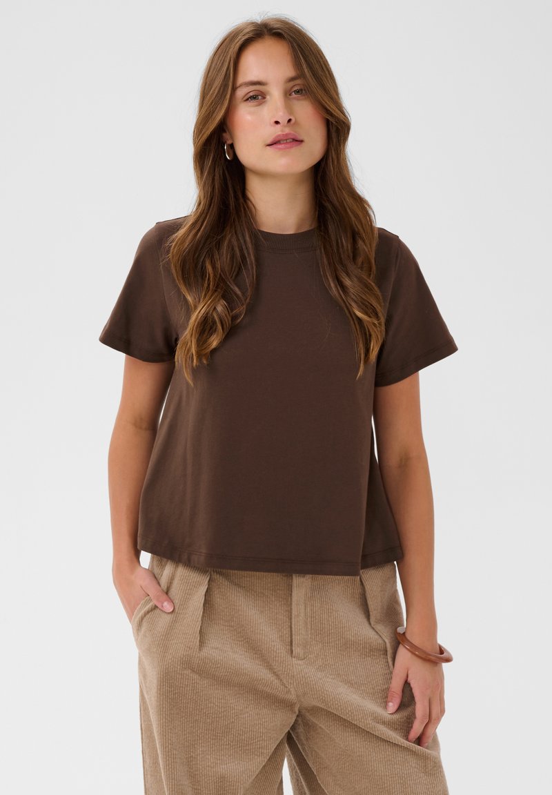 Femme aux longs cheveux bruns, portant un t-shirt marron foncé à manches courtes et un pantalon beige, debout les mains dans les poches devant un fond uni.