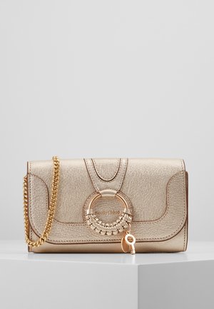 Pochette en cuir doré brillant avec anneau tressé et breloque clé, bandoulière chaîne dorée et accents de panneaux cousus sur un socle blanc.