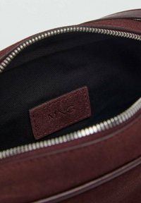 Intérieur d'un sac en daim bordeaux avec une doublure noire, une fermeture éclair en argent et une petite étiquette en cuir embossée avec "MNG" visible.