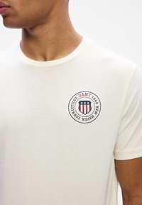 Lysecreme kortærmet t-shirt i bomuldsblanding, med et rundt logo med rød og blå skjold og tekstdetaljer. Enkel crew neck design.