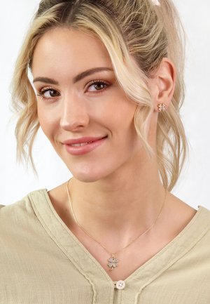 Goldfarbener Schmuck mit einem floralen Anhänger und glitzernden Akzenten, getragen zu einer hellgrünen, strukturierten Bluse. Einfaches, elegantes Design.