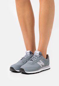 Scarpe sportive grigie con una parte superiore in sintetico liscio, dettagli bianchi, lacci testurizzati e una suola in gomma nera. Presentano il grande logo "N" sul lato.