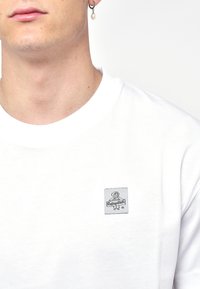 T-shirt bianco in cotone con colletto rotondo, caratterizzato da una piccola toppa di tessuto grigia con grafica e testo cucita sul lato sinistro.