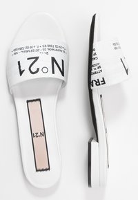 White slide sandals with wide straps featuring printed black text. Flat design with cushioned insole and subtle black accent on the sole.