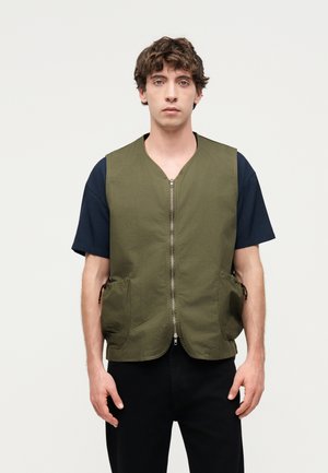 Junger Mann mit lockigem Haar, der eine olivgrüne Reißverschlussweste über einem schwarzen Kurzarmshirt trägt vor einem einfarbigen hellen Hintergrund.