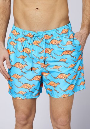 Mann trägt leuchtend blaue Badehose mit orangefarbenen Känguru-Mustern und gelben Punkten, Hände ruhen nahe der Hüften vor einem einfarbigen Hintergrund.