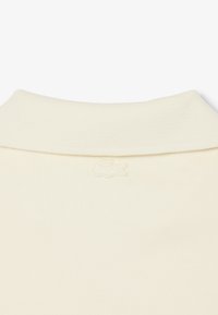 Cuello de punto acanalado color blanco roto con un sutil logo de cocodrilo bordado, centrado justo debajo del cuello.