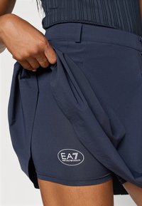 Temně modré sportovní šortky s hladkou texturou. Na spodním lemu je bílá logo EA7. Ruka tahá za látku, aby ukázala flexibilitu.