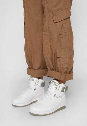 Białe wysokie trampki z beżowymi akcentami i wyrazistymi sznurówkami, noszone z podwiniętymi brązowymi spodniami cargo, ukazującymi szczegóły kieszeni.