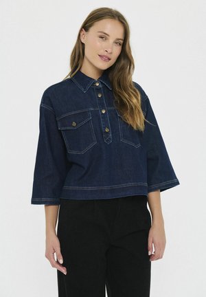 Frau mit langen Haaren, die eine dunkelblaue Jeansjacke mit großen Taschen und schwarze Hosen trägt, steht vor einem einfarbigen Hintergrund.