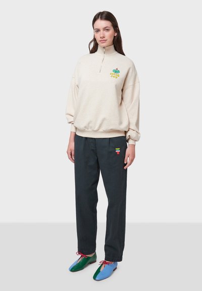 Felpa beige con zip e ricami verde e giallo, abbinata a pantaloni scuri con una striscia orizzontale multicolore. Scarpe multicolori.