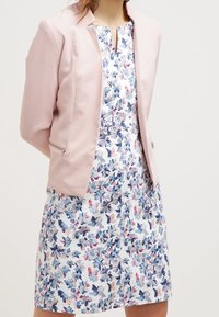 Lichtroze getextureerd jasje met ritszakken over een witte jurk met een blauw- en roze bloemenpatroon, korte mouwen en een V-hals.