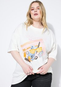 Hvid oversized t-shirt med et farverigt grafisk print af ferietema genstande, herunder solbriller, parfume og tilbehør.