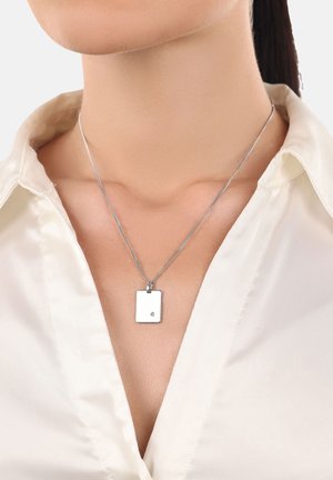 Schlüsselbeinsicht einer Person, die ein weißes Satinhemd und eine silberne rechteckige Anhängerkette trägt, mit einem kleinen Edelstein in der unteren rechten Ecke.