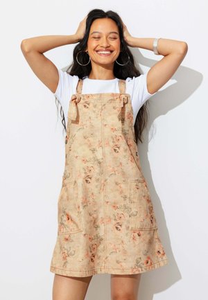 Femme souriante aux longs cheveux foncés porte une robe tablier beige fleurie par-dessus une chemise blanche, les mains derrière la tête sur fond blanc.
