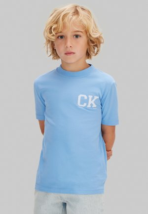 Ragazzo biondo con capelli ricci che indossa una maglietta CK azzurro chiaro e jeans chiari, in piedi davanti a uno sfondo grigio uniforme.