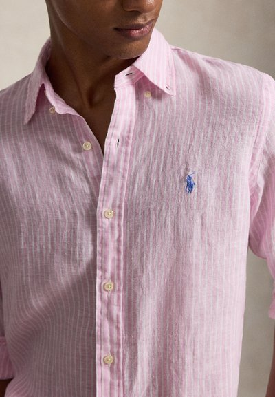 Mees kannab helesinine ja valge triibulist nööpidega särki, millel on sinine tikitud logo rinnal, näidatud kaelast vöökohani.