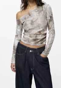 Langärmliges, schulterfreies Oberteil in metallischem Silber mit strukturiertem Stoff, kombiniert mit dunkelblauen Denim-Hosen mit sichtbaren Nähten.