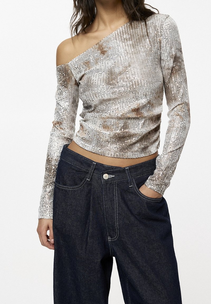 Langärmliges, schulterfreies Oberteil in metallischem Silber mit strukturiertem Stoff, kombiniert mit dunkelblauen Denim-Hosen mit sichtbaren Nähten.
