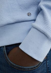 Világoskék, texturált pulóver, bordás mandzsettával és alsó résszel, kis ezüst emblémával. Sötétebb farmerrel párosítva, amit egy kéz tart a zsebben.
