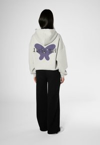Lys grå oversized hettegenser med et lilla sommerfugl-design og teksten "Lucky Girl" på ryggen. Kombinert med svarte vide bukser.