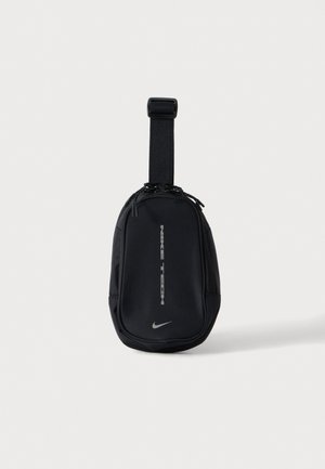 Čierne kompaktné puzdro Nike Tech s nastaviteľným popruhom a dvojitým zipsom, zobrazené na rovnomernom svetlom pozadí.