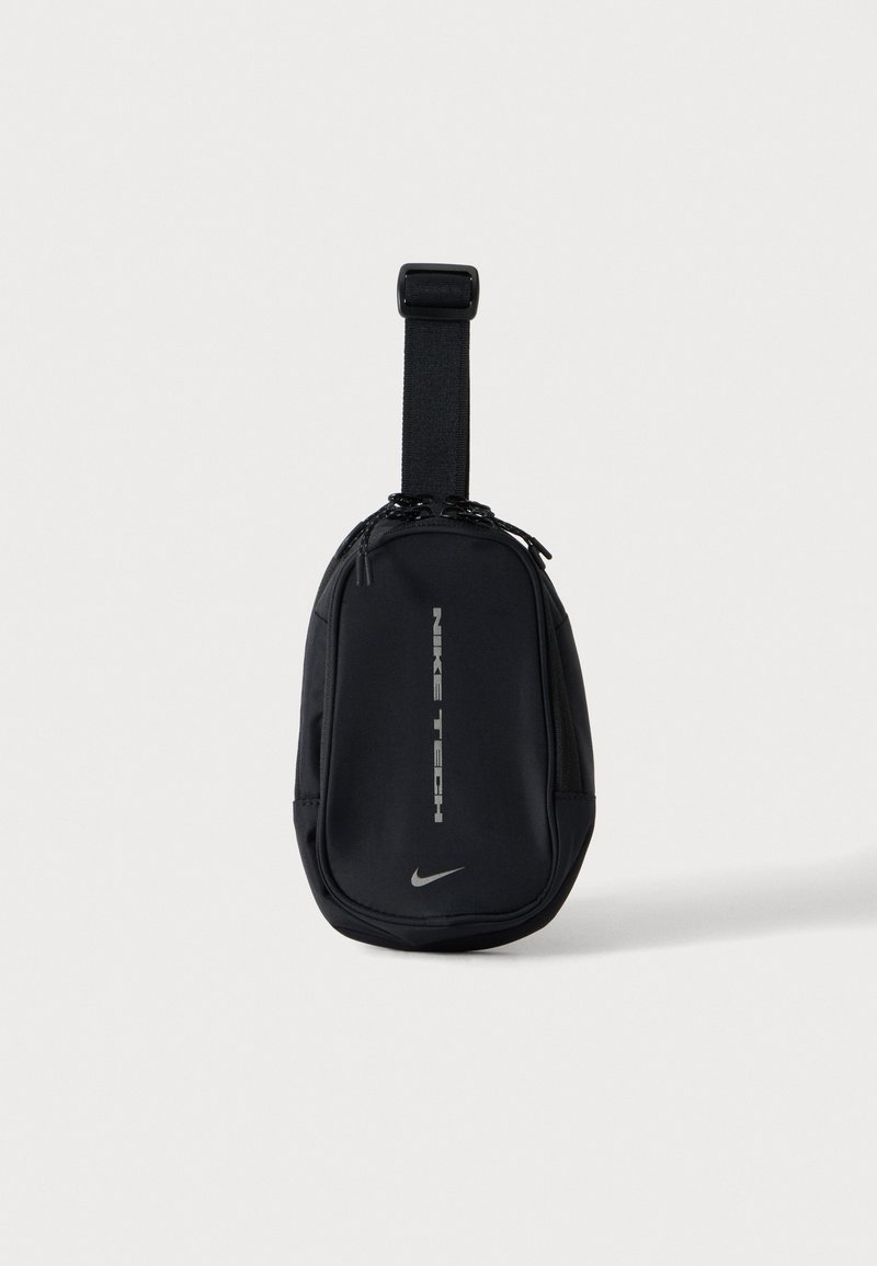 Sort kompakt Nike Tech taske med justerbar rem og dobbel lynlås, vist mod en ensartet lys baggrund.