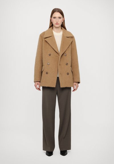 Brązowy wełniany płaszcz typu pea coat o teksturowanym wykończeniu, z podwójnymi guzikami, szerokimi klapami i dwoma przednimi kieszeniami, w połączeniu z kremowym swetrem i brązowymi spodniami.