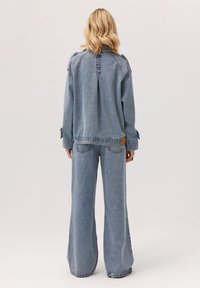 Osoba z włosami blond do ramion, ubrana w jasnoniebieską kurtkę dżinsową i szerokie jeansy, stojąca tyłem na jednolitym tle.