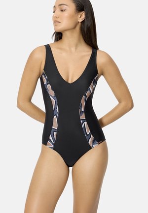 Mujer de pie con las manos detrás de la espalda, vistiendo un bañador de una pieza negro con paneles laterales de estampado geométrico.