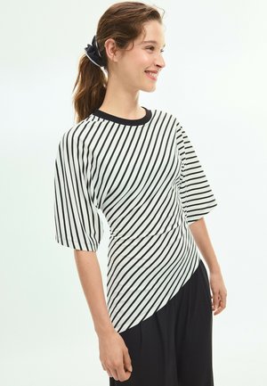 Jeune femme souriante portant un haut à manches courtes rayé en diagonale noir et blanc et un pantalon noir, avec les cheveux attachés en queue de cheval.