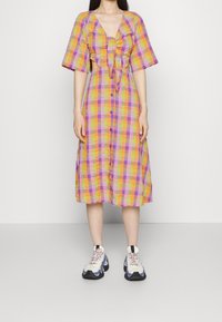 Robe à carreaux jaune, violette et orange, avec un col en V, des manches courtes, un nœud à l'avant et une fermeture boutonnée. Associée à des baskets épaisses.